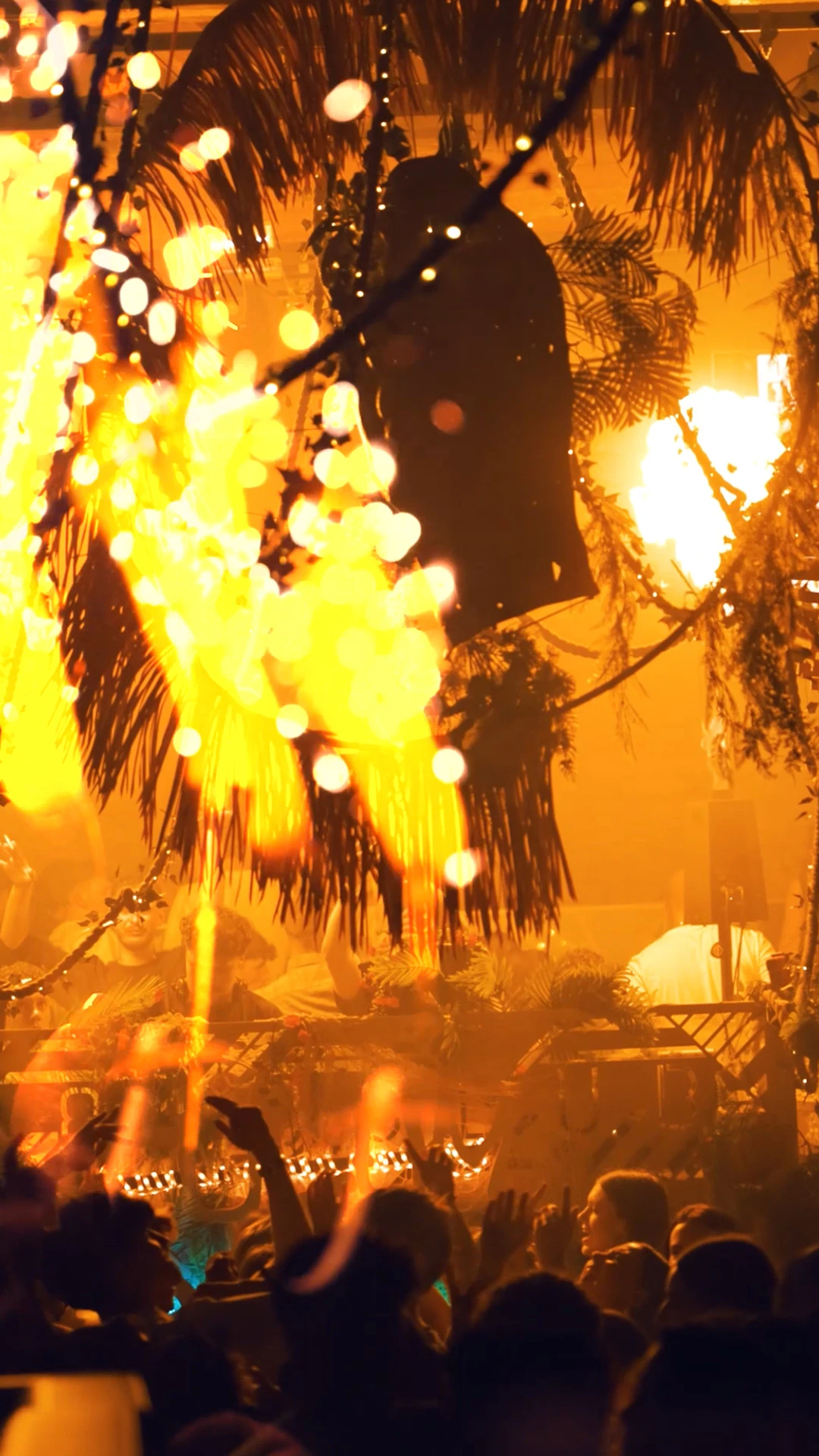 Foule dansante sous des lumières jaunes et orangées dans une ambiance tropicale avec feuillages et DJ en fond.