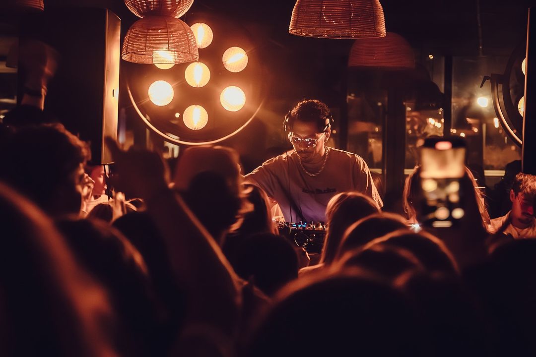 Un DJ mixant sur scène dans une ambiance chaleureuse, entouré d'une foule énergique sous des lumières tamisées et des suspensions décoratives.