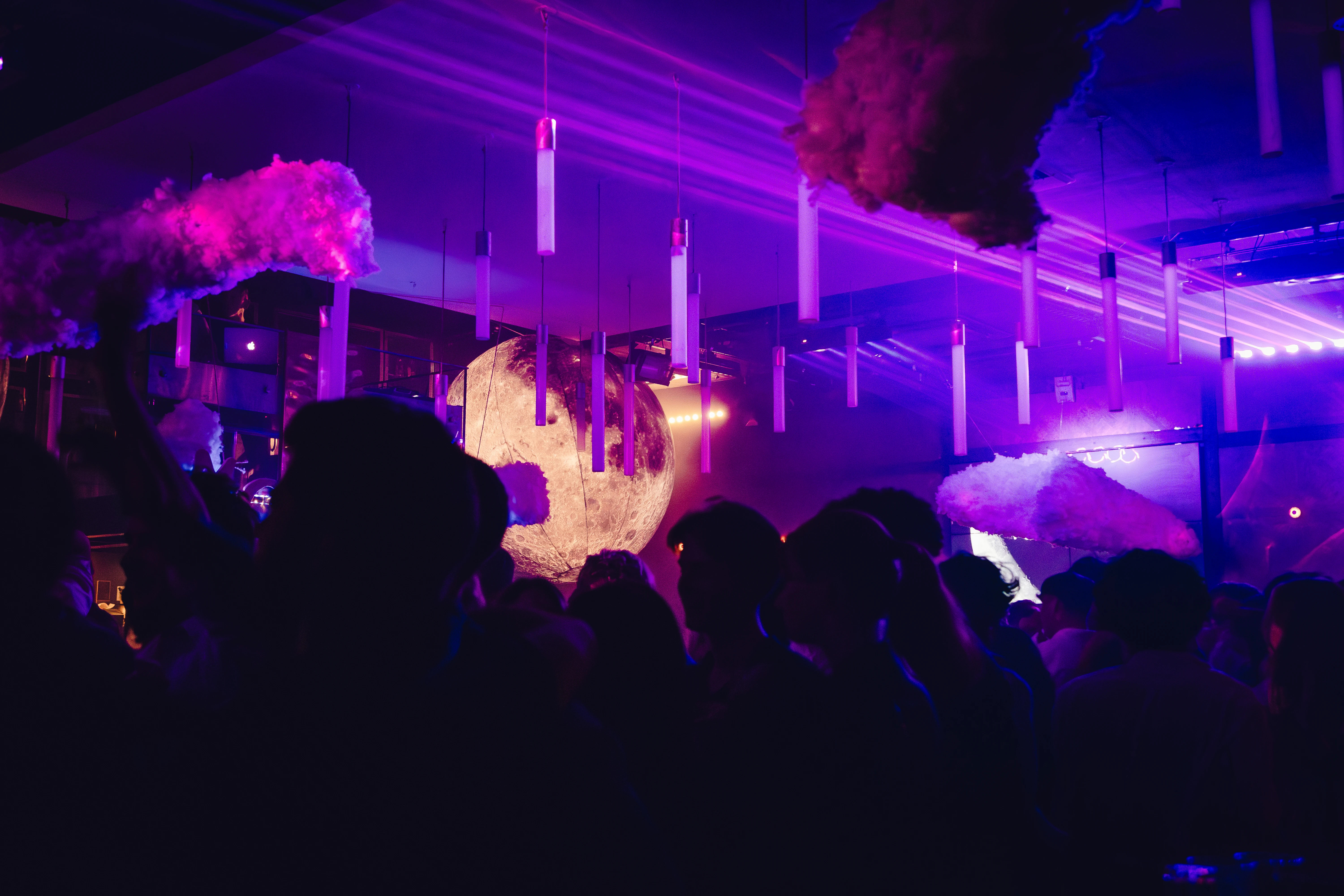 Foule de clubbers sous des lumières violettes et éléments suspendus en forme de nuages, avec une lune géante en arrière-plan.