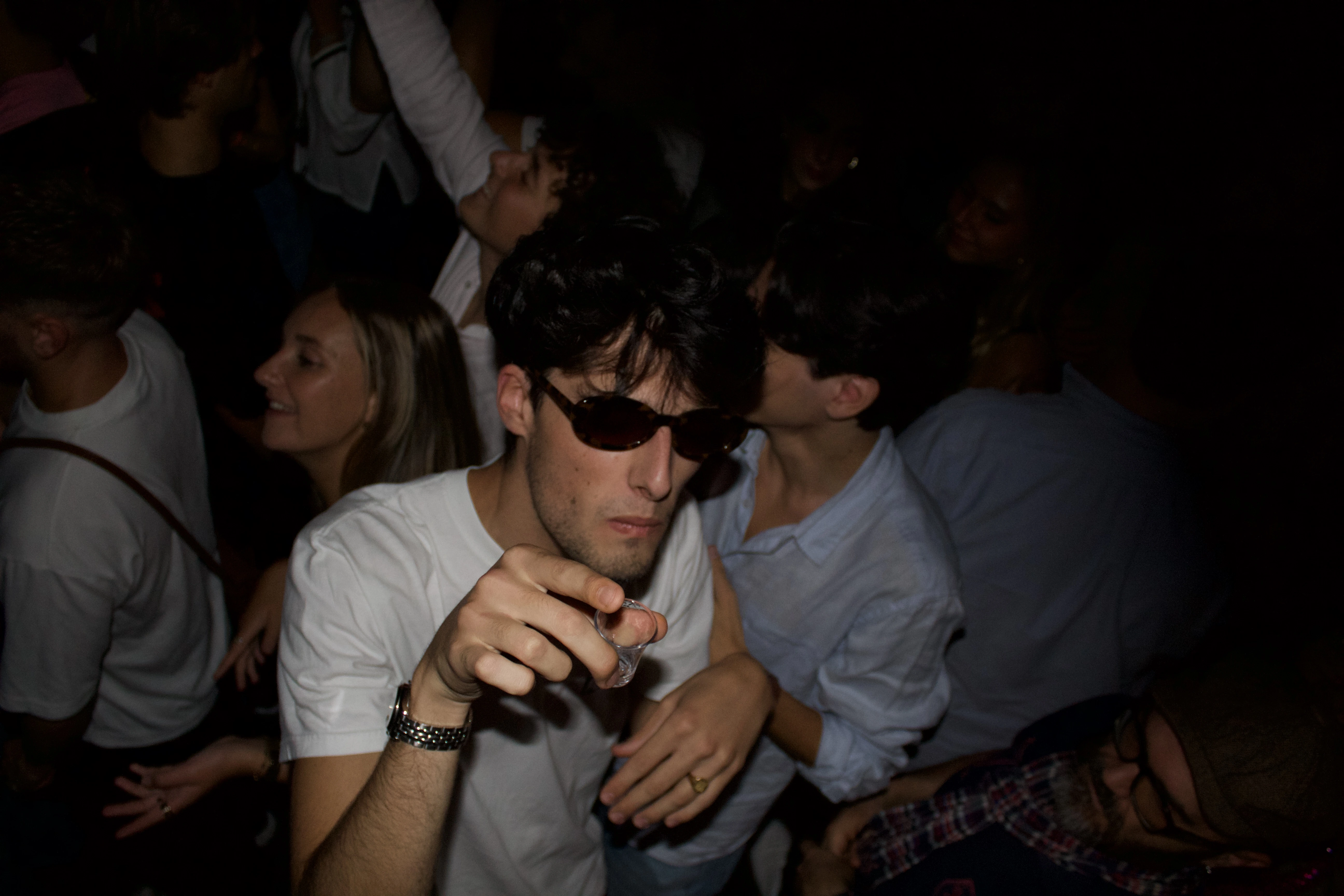Jeune homme vêtu de lunettes de soleil pointe la caméra avec un shot à la main.