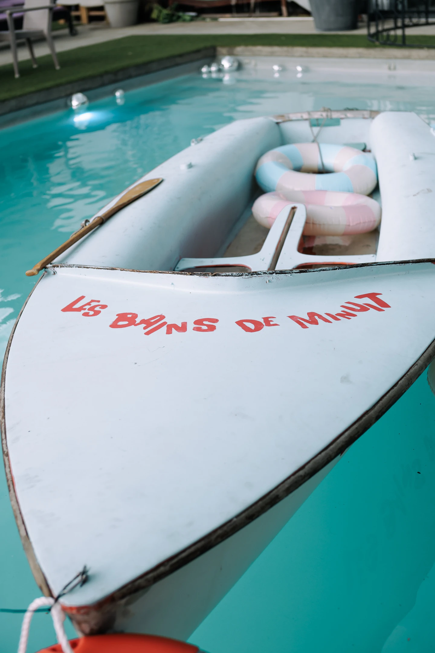 Bateau blanc avec une pagaie et deux bouées pastel, flottant dans une piscine, avec l’inscription Les Bains de Minuit sur la proue.