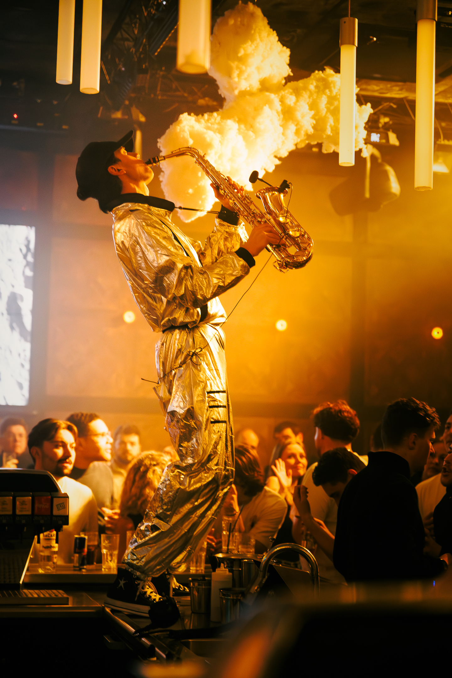 Homme en combinaison d'astronaute jouant du saxophone au milieu d'une foule de personnes le tout dans une ambiance chaleureuse