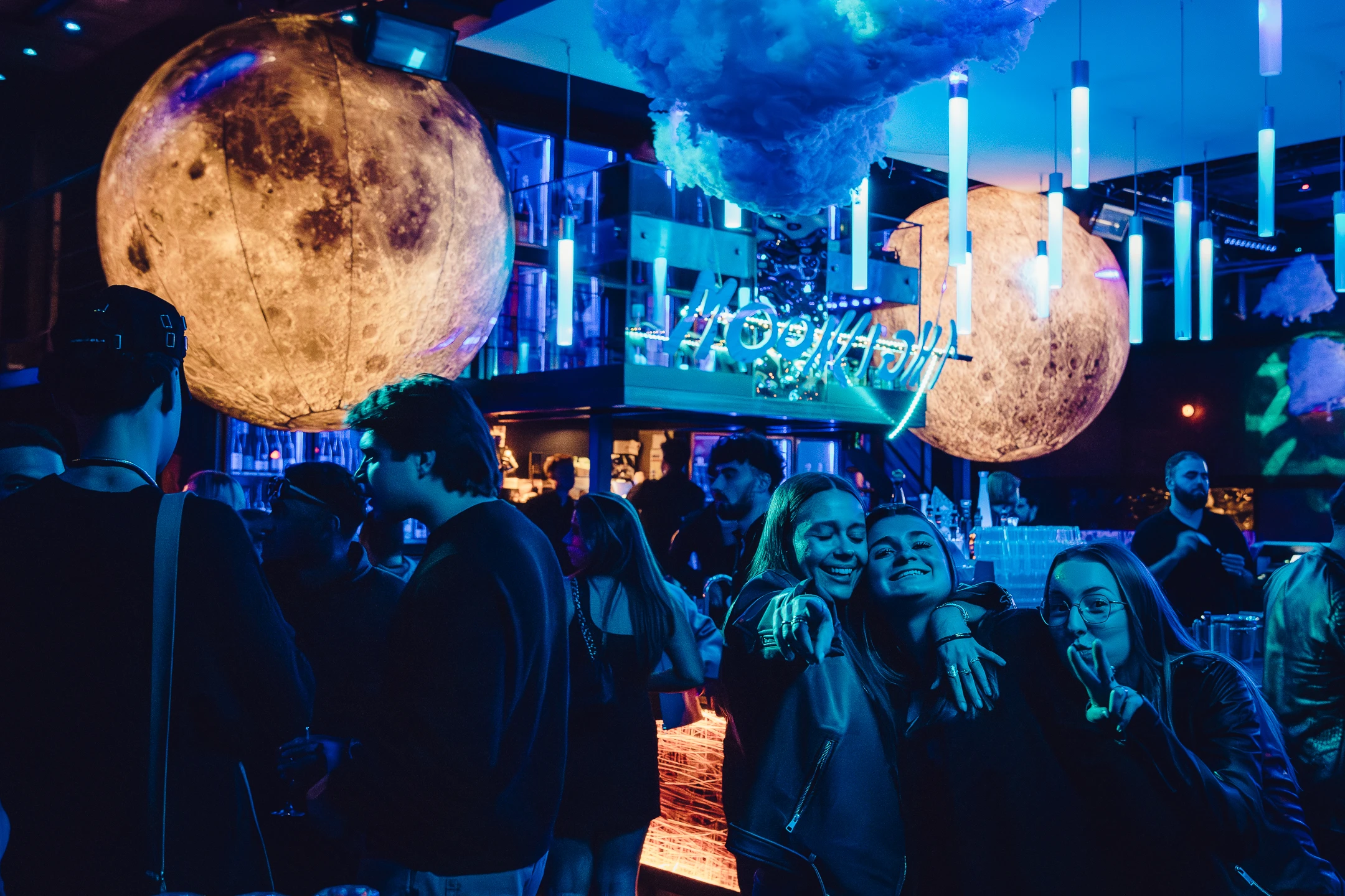 Ambiance de club futuriste avec des jeunes discutant sous des globes lunaires géants et des lumières bleues.
